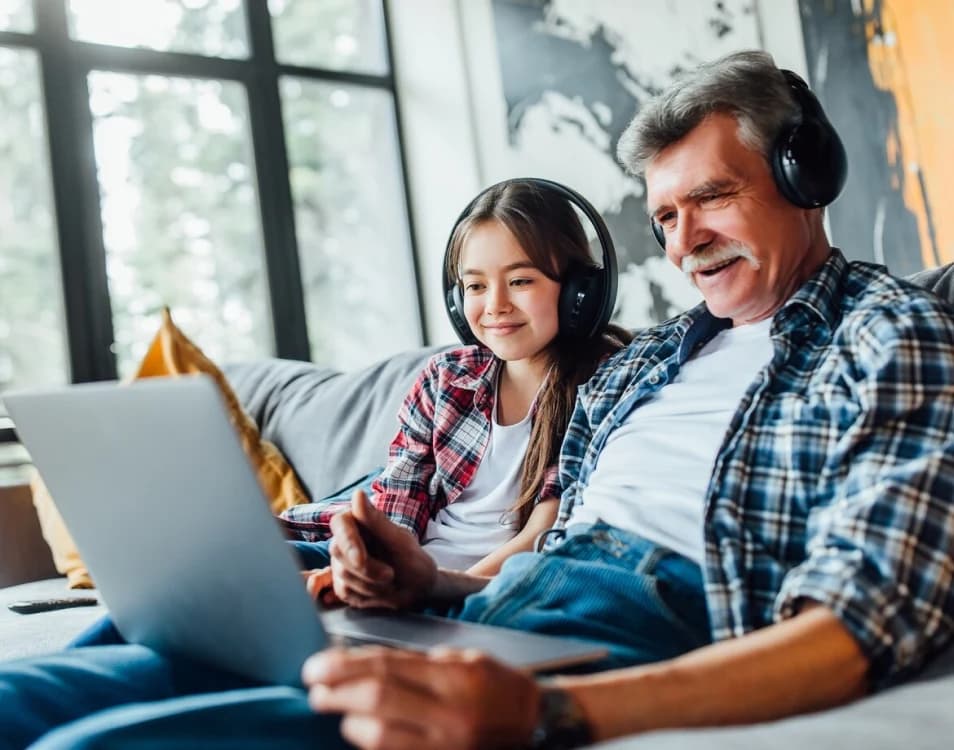 Grandfather and granddaughter on the sofa at home, wearing headphones, watching something together on a laptop